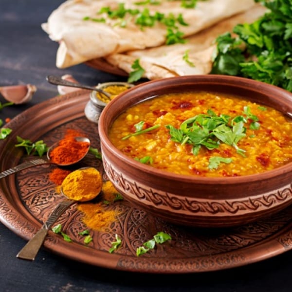 A bowl of lentil dal soup garnished with herbs, surrounded by spices, flatbread, and lentils on a copper tray.