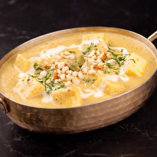 Creamy paneer curry in a brass bowl, garnished with nuts, herbs, and cardamom pods.