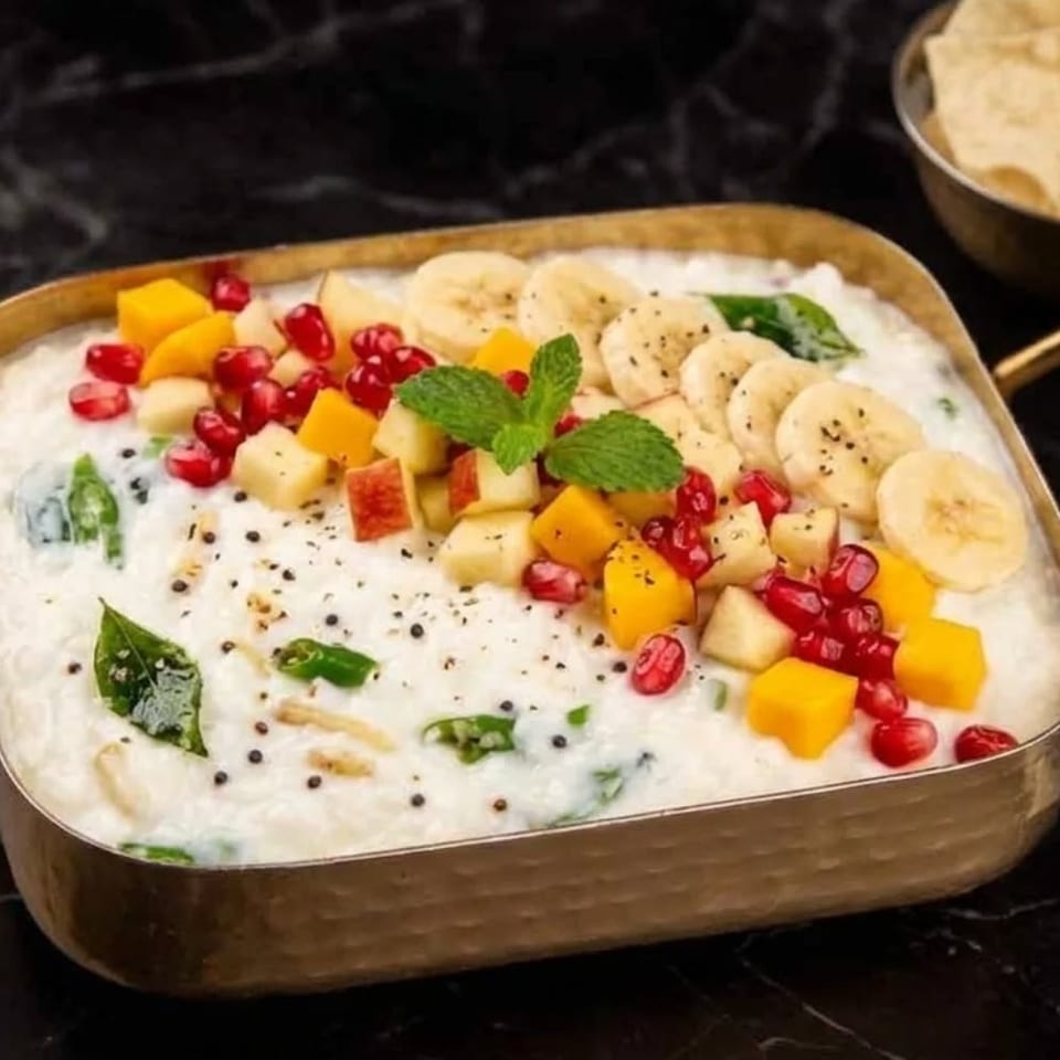 Creamy yogurt dish topped with colorful diced fruits, pomegranate seeds, and mint in a brass bowl.
