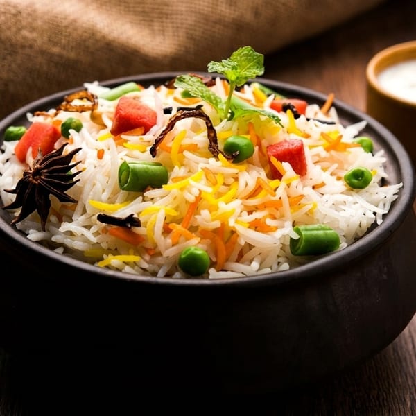 Colorful vegetable biryani rice in a black bowl with raita on a wooden table.