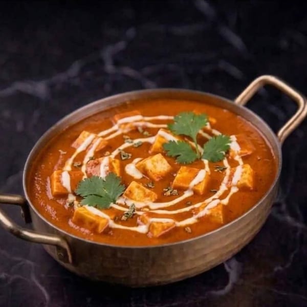 Paneer butter masala in a brass karahi, garnished with cream swirls and fresh cilantro.