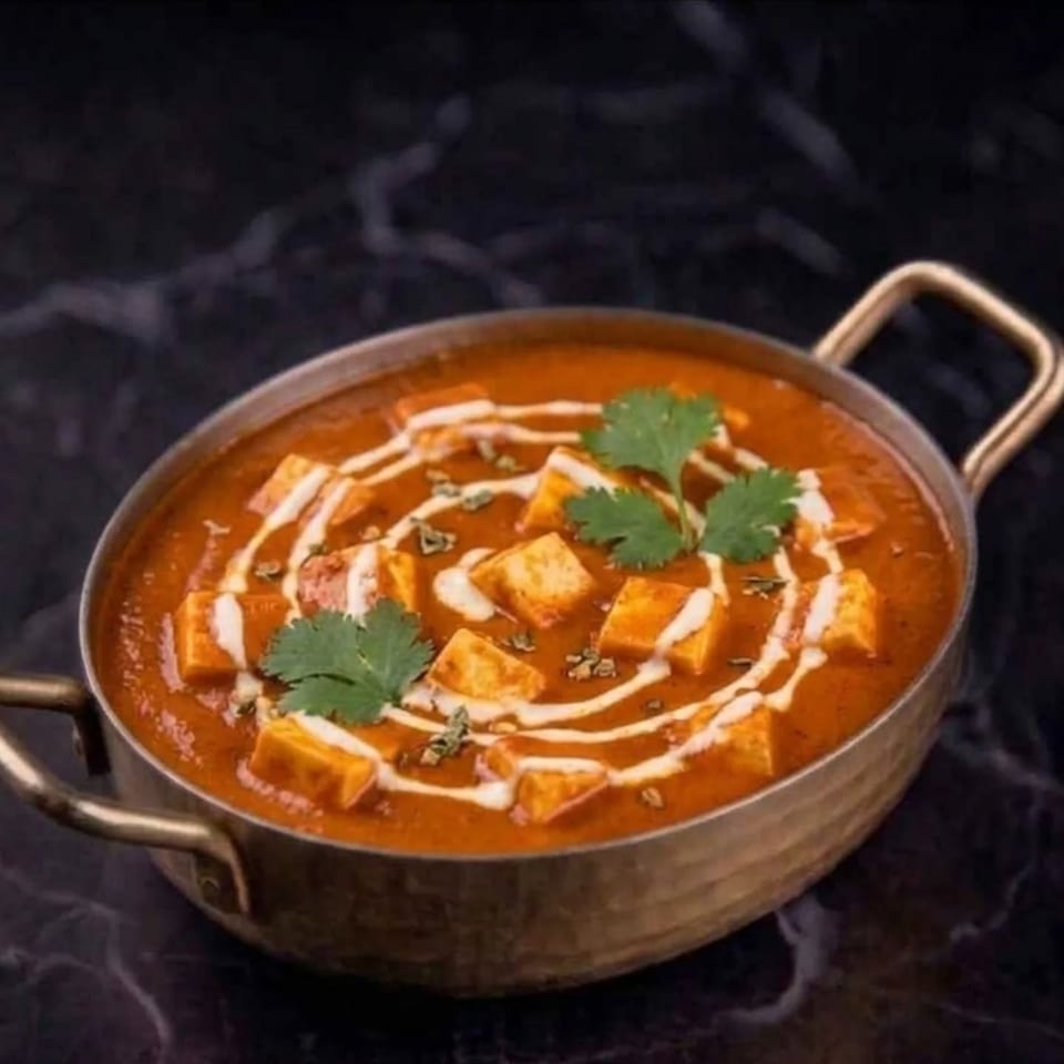 Paneer butter masala in a brass karahi, garnished with cream swirls and fresh cilantro.